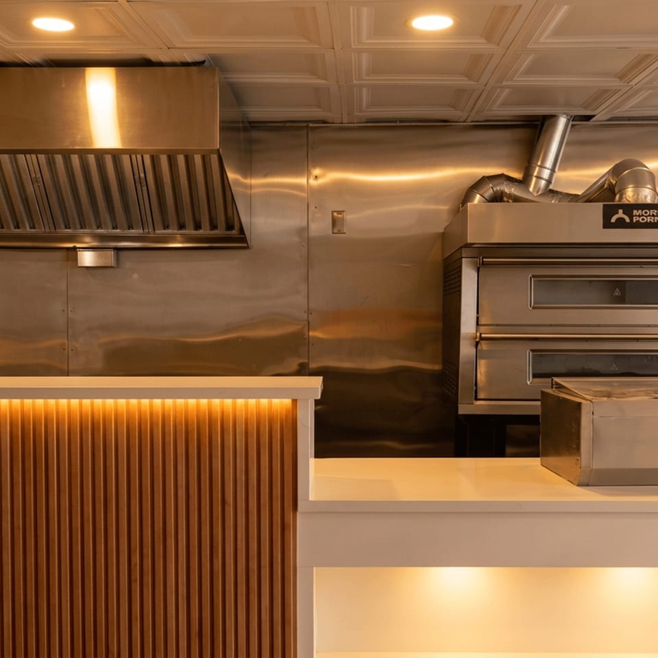 Warmly lit restaurant kitchen with stainless-steel ventilation hood, double ovens, countertop equipment, and decorative fluted wood paneling.